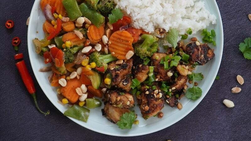 Chicken Kung Pao on cooked rice with sliced carrots and green vegetable on white ceramic plate